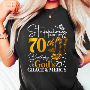 May include: Black t-shirt with white text that reads "Stepping into my 70th Birthday with God's Grace & Mercy". The number 70 is gold and there is a gold high heel shoe with glitter and a butterfly graphic.
