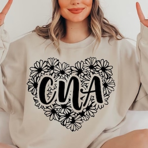 Puede incluir: Una sudadera beige con un diseño negro en forma de corazón que presenta las letras "CNA" rodeadas de flores de margarita estilizadas.