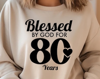May include: A beige sweatshirt with black text that reads "Blessed by God for 80 Years" with a small heart graphic.