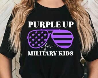Op de afbeelding: Zwart T-shirt met de tekst "PURPLE UP for MILITARY KIDS". Het ontwerp toont paarse zonnebrillen met sterren en strepen. Het T-shirt heeft een ronde hals en is in de taille geknoopt.