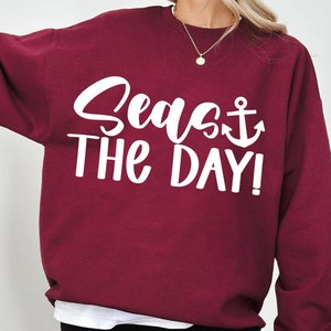 Könnte beinhalten: Burgunderfarbener Pullover mit weißem Text "Seas the Day!" und einem weißen Anker-Symbol.