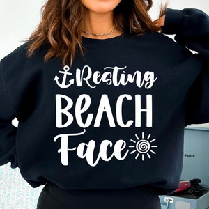 Könnte beinhalten: Schwarzer Pullover mit weißem Text, der "Resting Beach Face" mit einem Anker und einem Sonnenmotiv lautet.