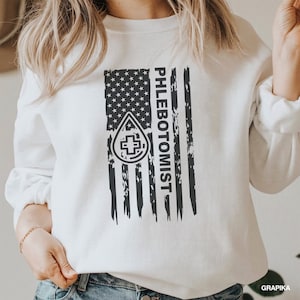 May include: White sweatshirt with a distressed American flag design. The flag has a black drop of blood with a white cross in the center and the word "PHLEBOTOMIST" written vertically on the right side.