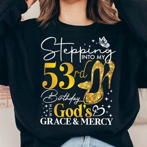 May include: Black sweatshirt with gold glitter text and graphic that reads "Stepping into my 53rd Birthday with God's Grace & Mercy". A gold glitter high heel shoe and a butterfly are part of the graphic.