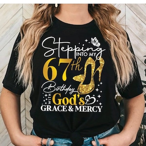 May include: Black t-shirt with gold glitter text that reads "Stepping into my 67th Birthday with God's Grace & Mercy". A gold glitter high heel shoe and a butterfly are also on the shirt.