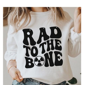 May include: White sweatshirt with black text that reads "Rad to the Bone" with a black radiation symbol in the middle of the word "Bone".