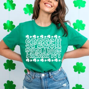 Op de afbeelding: Een groen T-shirt met witte tekst die "Speech Therapist" zegt, omringd door klavertjevier-ontwerpen.