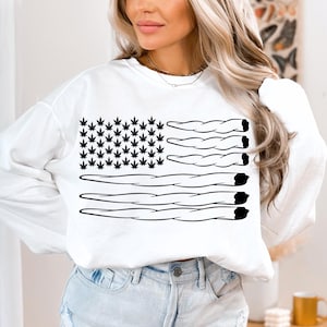 May include: White sweatshirt featuring a black and white graphic of an American flag made from marijuana leaves and joints.
