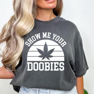 May include: A dark gray t-shirt with a white graphic of a marijuana leaf and the text "Show Me Your Doobie's" in a retro sunset design.