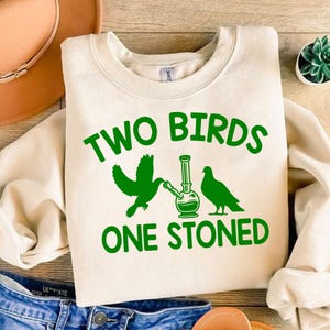 May include: A white sweatshirt with the text "Two Birds One Stoned" in green letters. The graphic features two birds, one holding a bong, and the other looking at it.