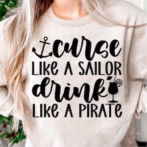 May include: A beige sweatshirt with the text "I curse like a sailor drink like a pirate" in black lettering. An anchor symbol is above the text and a cocktail glass with a lime wedge is to the right of the text.