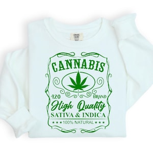 Könnte beinhalten: Weißes Sweatshirt mit einem grünen Aufdruck, der das Wort "CANNABIS" über einem Cannabisblatt zeigt. Darunter steht "High Quality Sativa & Indica 100% Natural". Das Sweatshirt hat lange Ärmel.