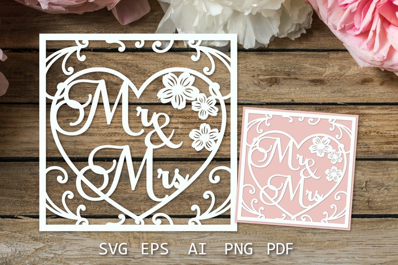 以下が含まれることがあります： 「Mr & Mrs」の文字が書かれたハート型の白い紙切り抜き。ハートは、花柄の装飾的な枠で囲まれています。デザインは木製の背景です。