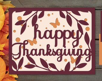 Pode incluir: Um cartão de papel recortado em 3D com várias camadas, com um fundo bordô e um interior creme. O cartão apresenta um design de videira com folhas e borboletas, e o texto "happy Thanksgiving".