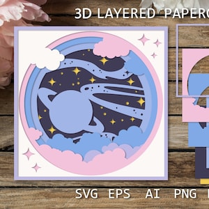 Könnte beinhalten: Ein 3D-Schnittmuster aus Papier mit einem Planeten mit Ringen, Wolken und Sternen in einem runden Rahmen. Das Design ist in Blautönen, Rosa und Gelb gehalten.