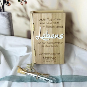 Könnte beinhalten: Eine Holzkiste mit dem Wort "Lebens" in weißer Schrift. Die Kiste enthält deutschen Text, darunter "Zur Kommunion von Mattheo 12. Mai 2026." Ein Glasröhrchen mit dem Wort "wunscherfueller" ist mit Bindfaden und Blumen gebunden.
