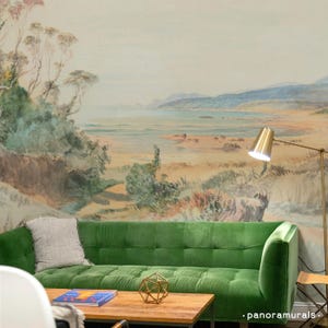 May include: A green velvet sofa with a light grey throw pillow sits in front of a watercolour mural depicting a coastal landscape with a body of water, hills, and trees. A gold lamp with a white shade is in the background.