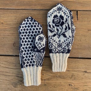 Può includere: Un paio di guanti lavorati a maglia con base color crema e motivi geometrici e floreali blu navy. I polsini sono color crema e i guanti sono esposti su una superficie di legno. Questi accessori invernali sono progettati per calore e stile.