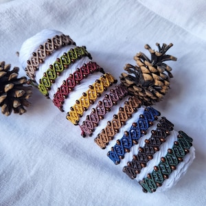 Puede incluir: Una colección de nueve pulseras de macramé en varios colores, incluyendo beige, verde, rojo, amarillo, morado, marrón, azul, marrón oscuro y verde oscuro, cada una con un patrón entrecruzado con pequeñas cuentas metálicas integradas.