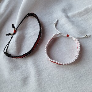 May include: Two friendship bracelets, one black and one white, with red accents. The bracelets are made with string and have small silver beads.