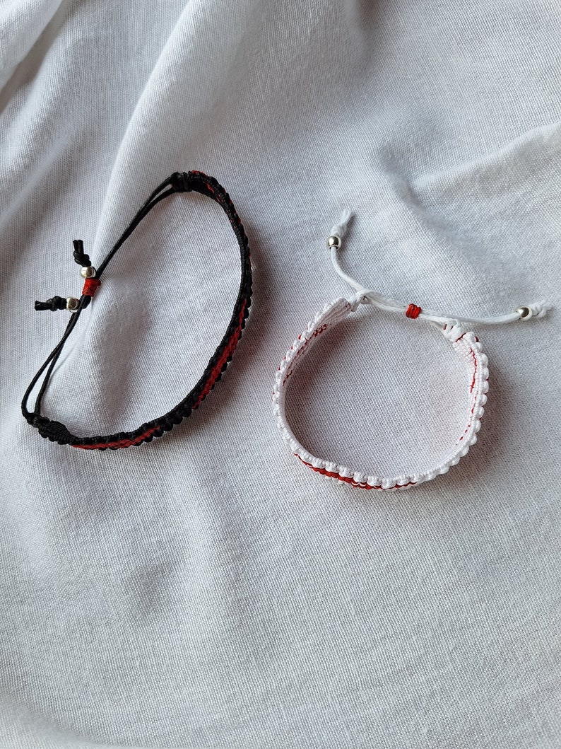 May include: Two friendship bracelets, one black and one white, with red accents. The bracelets are made with string and have small silver beads.