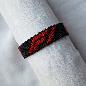 May include: A black and red friendship bracelet with a woven pattern. The bracelet is made of string and has a simple design.
