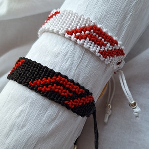 May include: Two friendship bracelets with red and white stripes and red and black stripes. The bracelets are tied with silver beads.