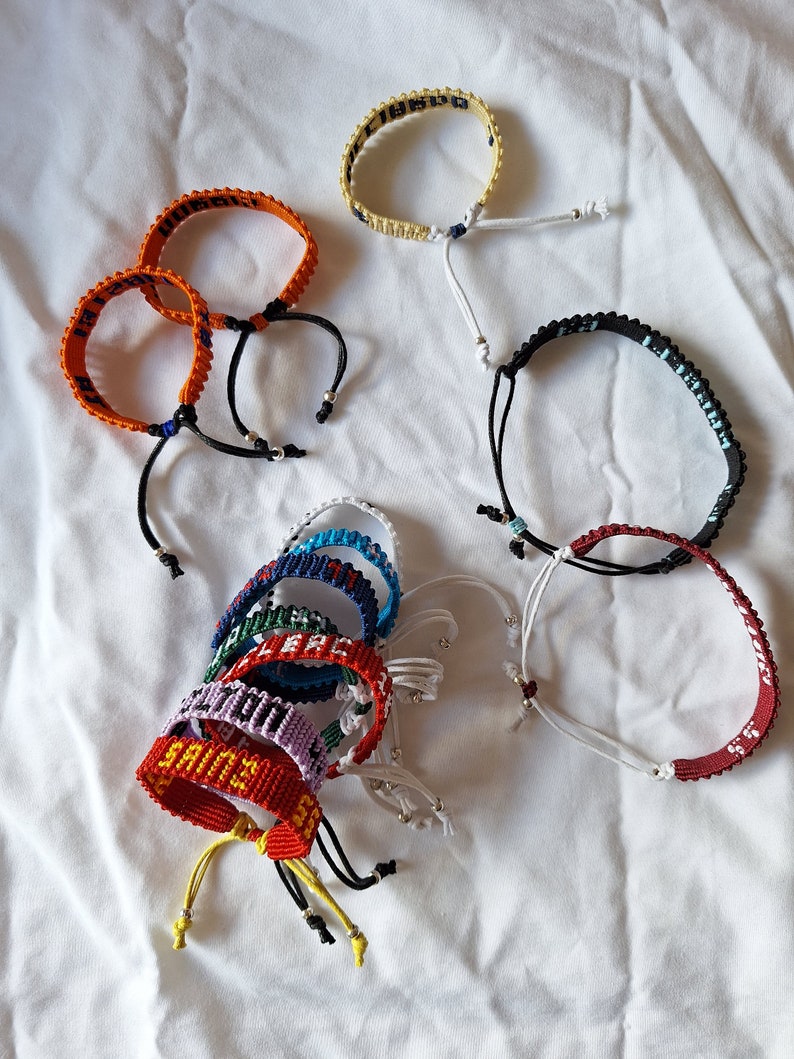May include: A collection of colorful friendship bracelets with various words and phrases woven into the design. The bracelets are made with colorful threads and have adjustable closures.
