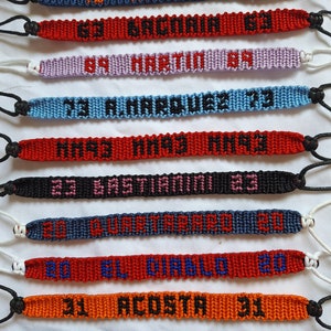 May include: Ten colorful friendship bracelets with black and orange lettering. The bracelets are made with woven string and feature names and numbers. The bracelets are arranged vertically, with the top bracelet being blue and the bottom bracelet being black.