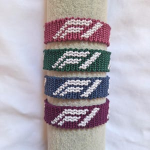 May include: Four woven friendship bracelets arranged vertically. Each bracelet features a white geometric logo pattern against a solid background color: dusty rose, forest green, slate blue, and deep plum. The bands are crafted with a tight knot texture.