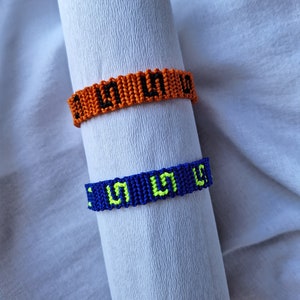 May include: Two friendship bracelets with black lettering on a white background. The top bracelet is orange with black lettering and the bottom bracelet is blue with yellow lettering.