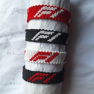 May include: Four friendship bracelets stacked on top of each other. The bracelets are made with red, white, and black string. The bracelets have a pattern of a white wave on a red background, a white wave on a black background, a red wave on a white background, and a red wave on a black background.