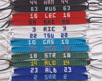 Könnte beinhalten: Eine Sammlung gewebter Freundschaftsarmbänder in verschiedenen Farben, die jeweils einen Namen oder Initialen und eine Nummer zeigen. Die Armbänder sind in Blau-, Orange-, Schwarz-, Rot-, Weiß-, Türkis-, Grün- und Blautönen gehalten. Jedes Armband hat einen geknoteten Kordelverschluss.