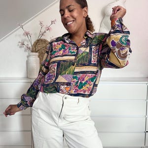 Puede incluir: Una camisa de manga larga con botones y un estampado geométrico y floral abstracto multicolor en tonos morado, verde, rosa y dorado, combinada con pantalones de pana blancos de talle alto con cierre de cremallera y botón.