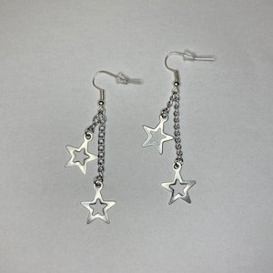 May include: A pair of silver dangle earrings with three star charms on each earring. The stars have a cut-out design.
