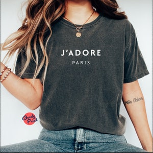 May include: A dark gray t-shirt with the text "J'ADORE PARIS" printed in white on the front. The shirt is made of a soft, comfortable material and has a relaxed fit.