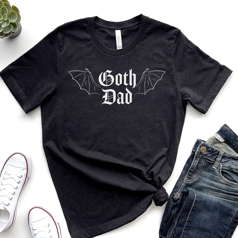 May include: Dark grey t-shirt featuring the words "Goth Dad" in a white, gothic font, with bat wings. The shirt is styled with blue jeans and white trainers, offering a casual, alternative aesthetic.