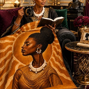 May include: A rectangular throw blanket featuring a detailed illustration of a person with a high bun hairstyle, wearing a white shell necklace and gold hoop earrings, set against a golden sunburst pattern with wavy lines in shades of amber and brown.