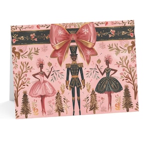 May include: A pink Christmas card featuring a black and gold Nutcracker and two ballerinas. The Nutcracker is holding a sprig of holly and the ballerinas are wearing pink and green tutus. A large red bow with gold snowflakes is at the top of the card.
