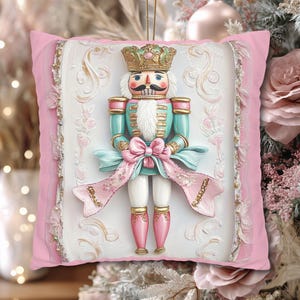 May include: A decorative Christmas ornament featuring a nutcracker soldier design. The ornament is set on a pink pillow-shaped background with a white embossed panel. The nutcracker is in teal, pink, and gold, with a large pink bow and a gold crown. A gold string is attached for hanging.