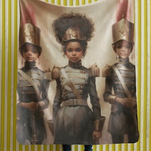 May include: A beige fleece throw blanket with a painting of three girls in military uniforms. The girls are wearing hats with gold trim and have dark brown hair. The painting is in a realistic style and has a vintage feel.