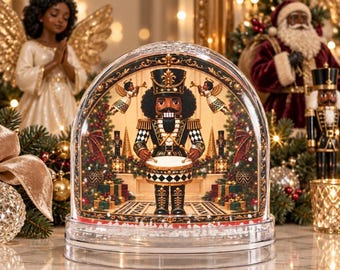 May include: A clear, arched glass snow globe featuring a central nutcracker figure in a black and gold uniform playing a drum. The scene includes two small angels with trumpets, miniature nutcrackers, and wrapped gifts on a tiled floor.