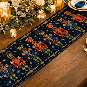 May include: A rectangular table runner featuring a deep navy blue background with a repeating pattern of nutcracker figures in red, green, and gold uniforms, interspersed with small gold stars and a decorative gold border along the edges.