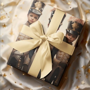 May include: A wrapped gift box featuring an image of a young person in a military-style uniform and hat. The box is tied with a wide, light gold satin ribbon and bow. The background is a cream-colored fabric with gold star accents.