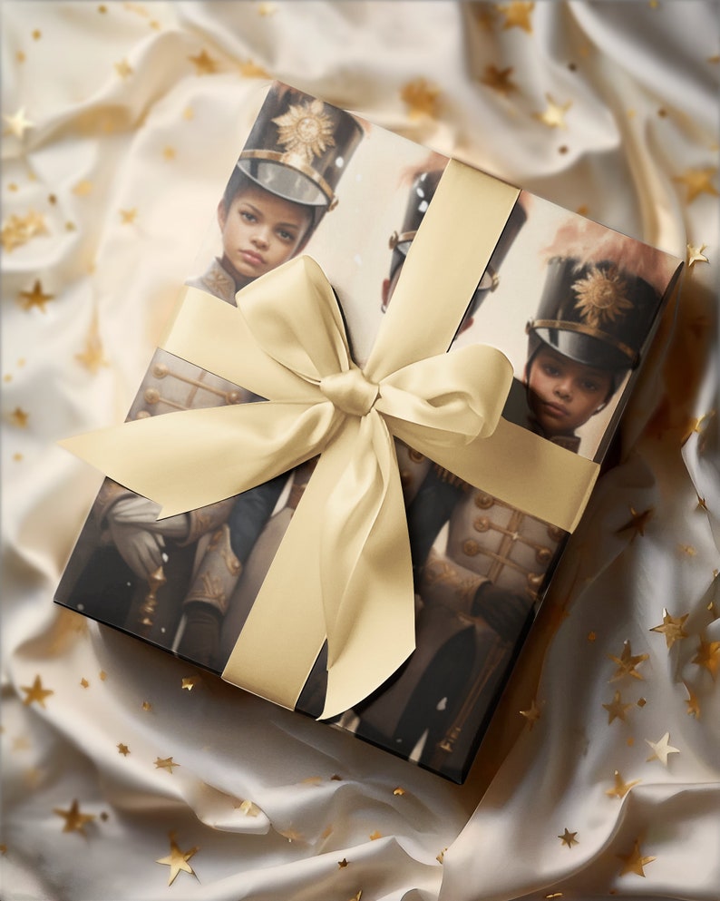 May include: A wrapped gift box featuring an image of a young person in a military-style uniform and hat. The box is tied with a wide, light gold satin ribbon and bow. The background is a cream-colored fabric with gold star accents.