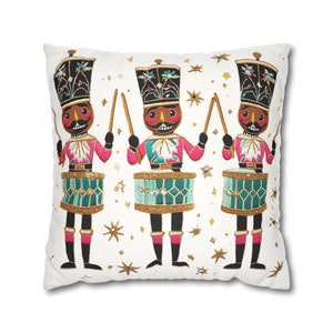 May include: White throw pillow with an embroidered design of three nutcracker soldiers playing drums. The soldiers are wearing red and blue uniforms with gold accents and black hats. The background is white with gold stars.