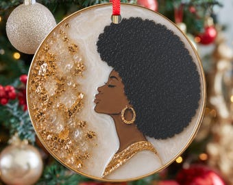 May include: Round Christmas ornament featuring a profile of a Black woman with a large afro, gold hoop earrings, and a gold necklace. The ornament has a gold border and a red ribbon for hanging. Gold glitter accents the design.