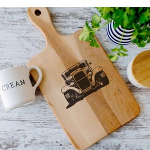 May include: Wooden cutting board with a vintage car design. The board is light brown with a handle and a dark brown illustration of a classic car. A white mug with the word "CREAM" is on the left.