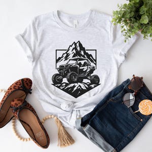 Peut inclure: Un t-shirt blanc avec un motif noir d'un v&eacute;hicule tout-terrain conduisant &agrave; travers une cha&icirc;ne de montagnes.