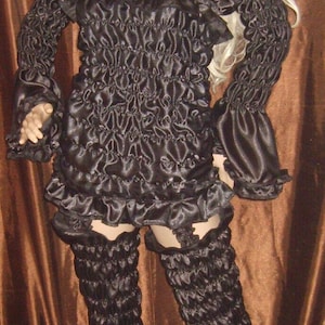 May include: Black satin costume with a ruffled top, long sleeves, and matching leg warmers. The outfit is displayed on a mannequin with a blonde wig, set against a brown backdrop.
